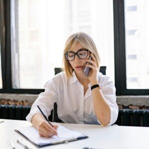 A woman in an office setting multitasking with a phone call and taking notes.